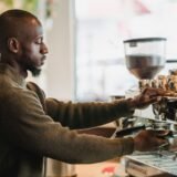 Interior de cafenea mică, cu un espressor pe tejghea, rafturi din lemn și câteva mese lângă o fereastră spre stradă