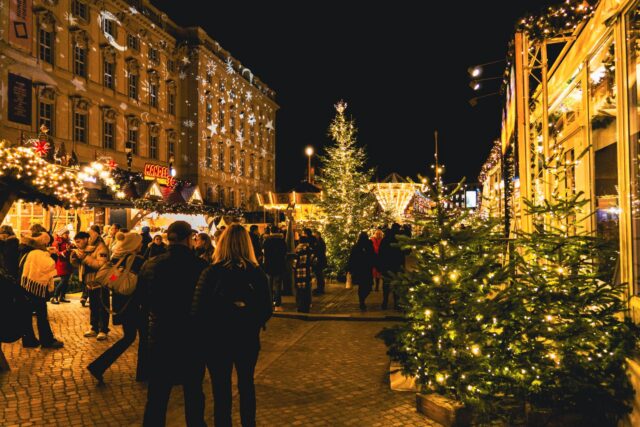 Căsuțe de lemn luminate ale unui târg de Crăciun într-o piață istorică, seara, cu un brad mare împodobit în centru